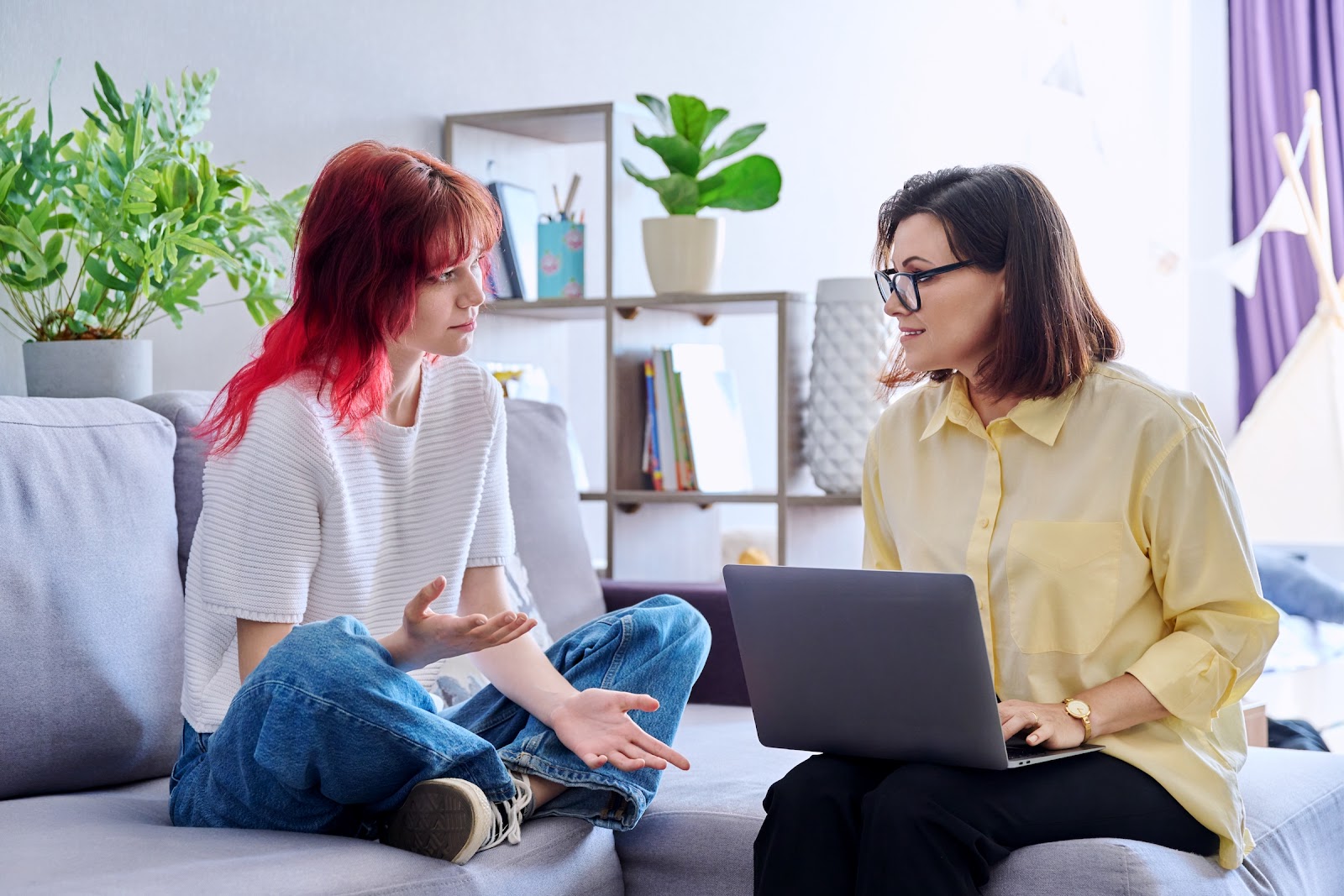 How to know if a therapist is right for you comes down to asking the right questions and knowing if you feel comfortable. In this image, a young woman sits cross-legged on a couch across from a therapist with a laptop. The girl's palms are facing up, as if she's telling a story.