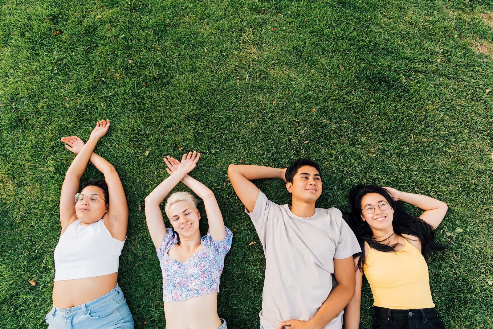 Practicing real self care is so important to maintaining your mental health. In this photo, a group of four friends lie on the green grass, arms above their heads, smiling.