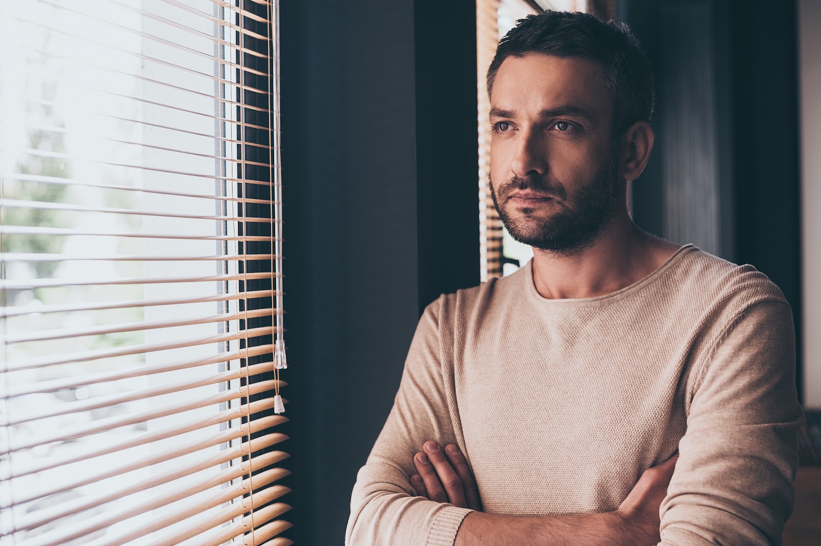 Negative self-talk gets in the way of practicing self-compassion. In this image, a dark-haired man stands in front of a dark wall, looking out a window. His expression is calm and thoughtful.