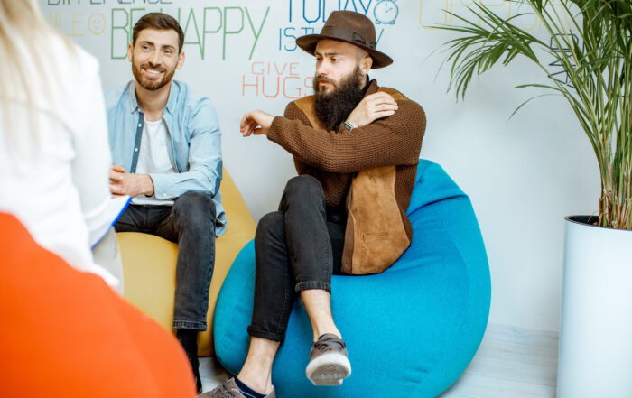 Working with an affirming therapist can help us feel comfortable expressing our true selves in a therapy setting. In this image, a couple sits across from a therapist. The man appears to be listening attentively to what his partner is telling the therapist.