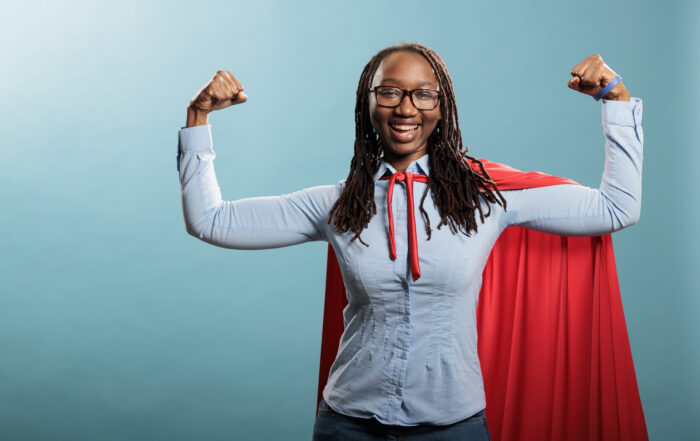 Happy positive justice defender flexing arms as a sign of strength while standing on blue background. Proud and tough looking young adult superhero woman showing empowerement and braveness.