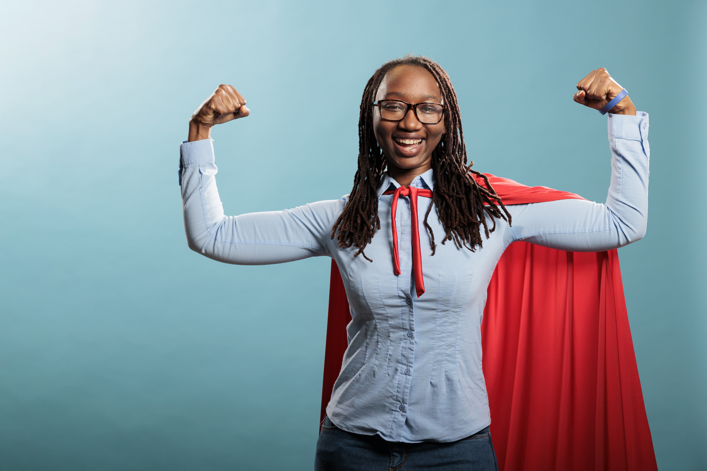 Happy positive justice defender flexing arms as a sign of strength while standing on blue background. Proud and tough looking young adult superhero woman showing empowerement and braveness.