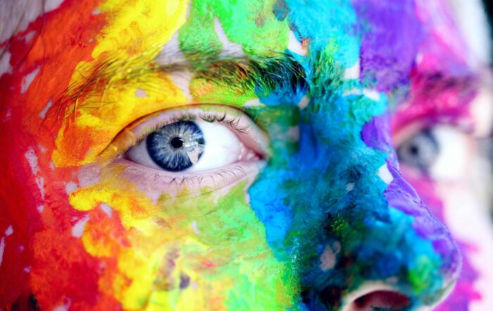 A close up of a person's eyes with their skin covered in face paint of the colors of the rainbow to celebrate LGBTQIA+ representation.