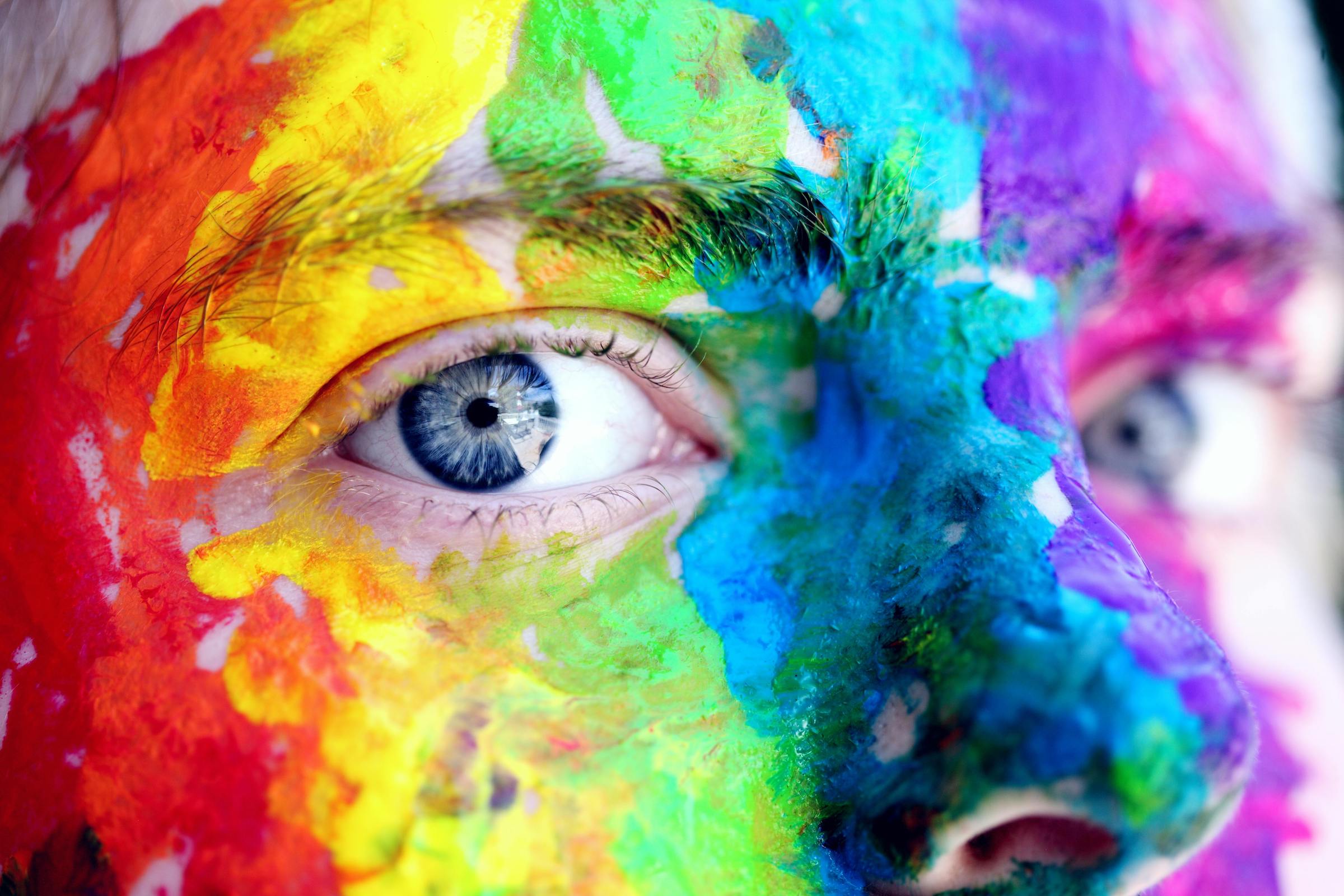 A close up of a person's eyes with their skin covered in face paint of the colors of the rainbow to celebrate LGBTQIA+ representation.